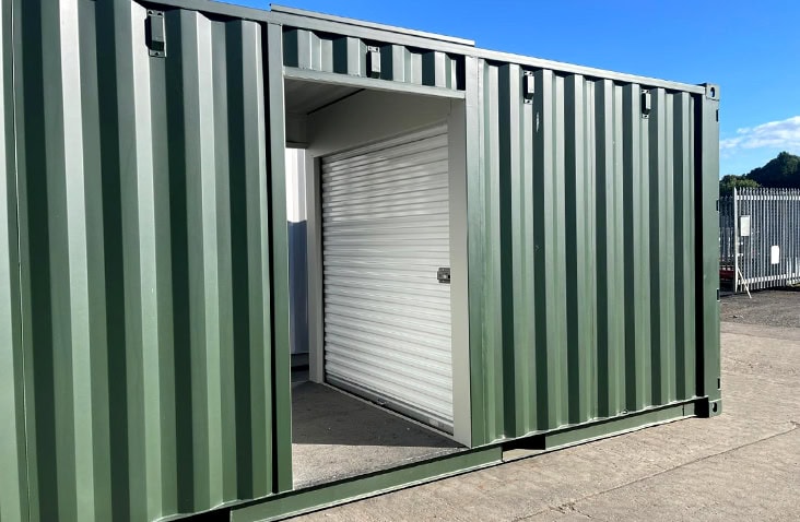 Green topper unit with a central doorway revealing a white roller shutter door inside. The container is placed on a concrete surface outdoors, with a metal fence and clear blue sky in the background. 