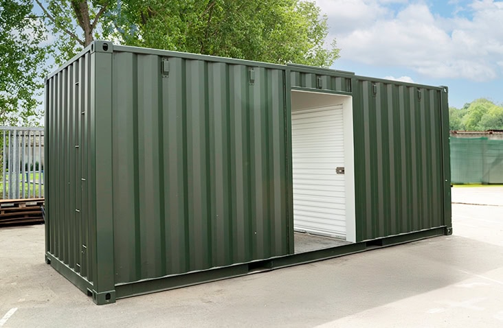 A green topper shipping container with a roller shutter door opening on the side, shown outdoors on a concrete surface with trees in the background. 