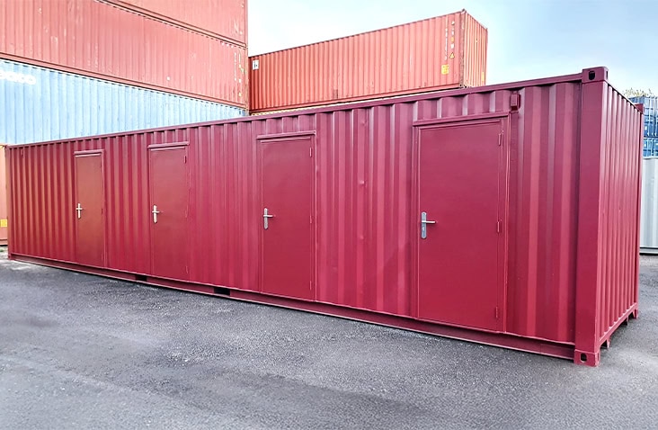 Burgundy multi-compartment storage container featuring four external personnel doors, photographed in a shipping container depot. 