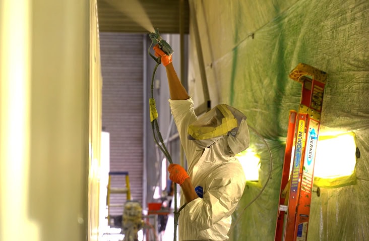 Technician in protective gear spray-painting a shipping container inside a controlled paint booth environment