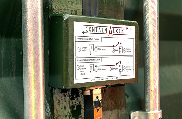 A Contain-A-Lock unit fitted to a green shipping container door, shown with a padlock inserted into the lock box mechanism. The printed operating instructions on the box are clearly visible, along with the container’s metal locking bars.