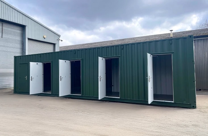 A long green multi-store container divided into five individual storage units, each with a white steel personnel door open to show internal space. 