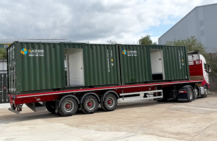 Two green steel shipping containers with side openings, branded with ‘S Jones Containers’ logo and contact number, loaded on a red flatbed trailer attached to a white Volvo truck. The scene is outdoors on a concrete surface near industrial buildings under a cloudy sky. 