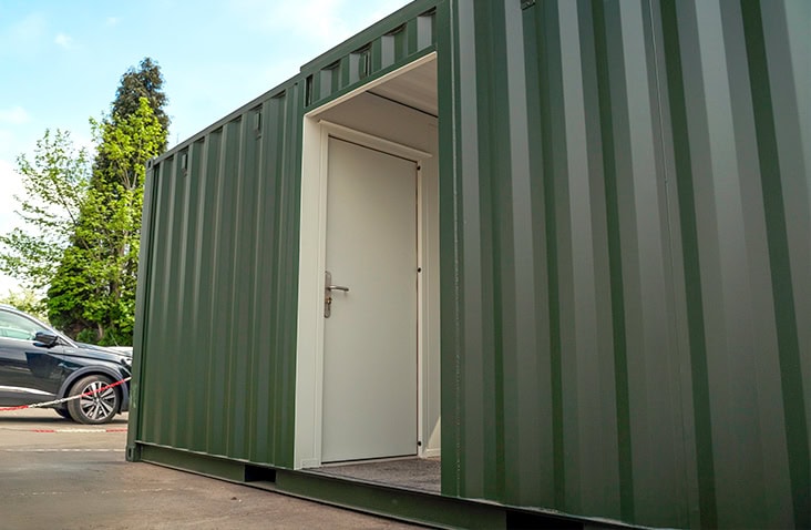 Green topper unit with a central doorway leading to a white personnel door fitted with a silver handle. The container is placed outdoors on a concrete surface, with a car and trees visible in the background under a partly cloudy sky. 