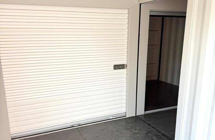 Interior view of a container unit showing a white roller shutter door on the left and an open doorway leading to another section of topper storage with dark flooring. 