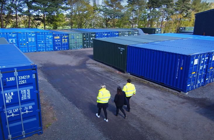 Three people wearing high-visibility jackets are walking through a self storage facility containing multiple blue and green shipping containers. 