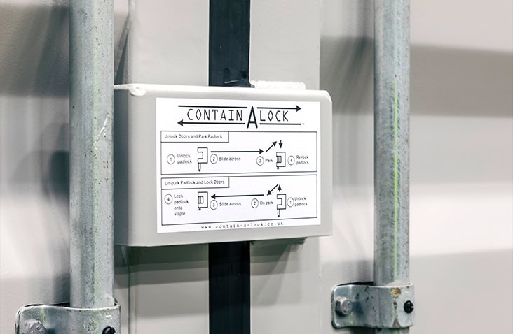A white Contain-A-Lock box installed on a light-coloured shipping container door. The instruction label is clearly displayed, and the surrounding vertical locking bars and fixings frame the lock box.
