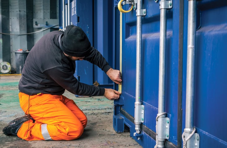     Engineer kneeling to measure and repair the seal area on a blue shipping container door. 