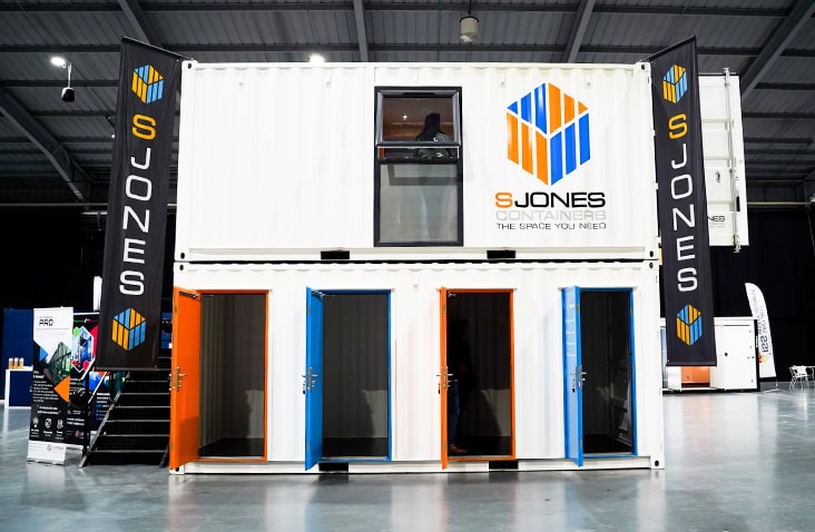 White two-storey exhibition container with a multi store unit on the ground level and branded S Jones graphics displayed at an indoor event. 