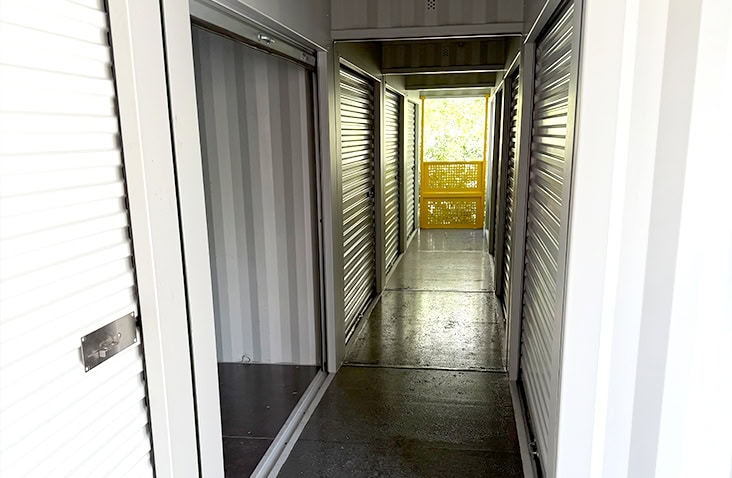 Interior corridor of a container unit with multiple roller shutter doors on both sides, leading to a bright yellow balustrade at the far end.