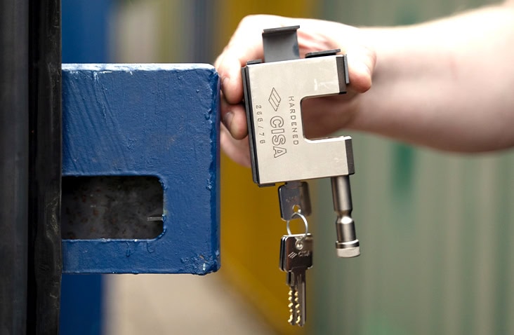 A close-up of a person holding a silver CISA hardened padlock with keys attached, positioned next to the blue steel locking box on a shipping container door. The padlock is shown aligned as if ready to be inserted into the lock housing, with the container’s coloured units blurred in the background.