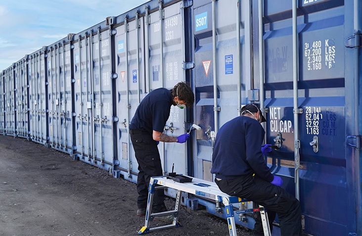 Two technicians restoring the exterior of blue shipping containers, applying ClearRestore® treatment to door fittings on a row of containers. 