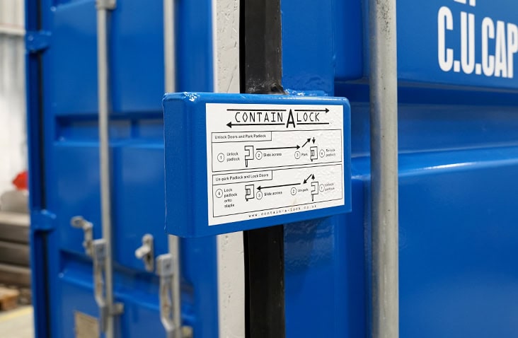 A blue Contain-A-Lock shroud fitted to a blue shipping container door, with an instruction label showing how to operate the locking system. The shroud is mounted over the door’s central locking bar, and the surrounding container doors and metal fixings are visible in the background inside an indoor workshop setting. 