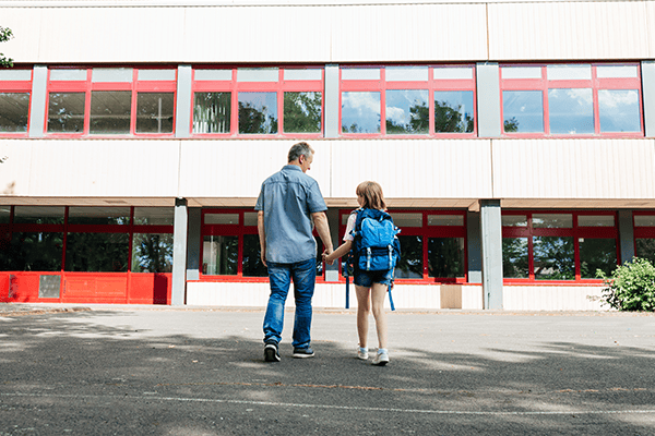 Adult and child walking hand in hand towards a modern school building, representing education, learning environments and student wellbeing. 