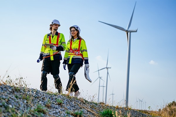 Two renewable energy engineers wearing safety gear walk near wind turbines at a wind farm, representing sustainable energy and renewables infrastructure. 