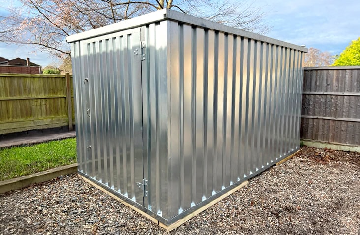 Galvanised 2m x 3m flat pack storage unit installed in a back garden on a gravel base, positioned against wooden fencing. 