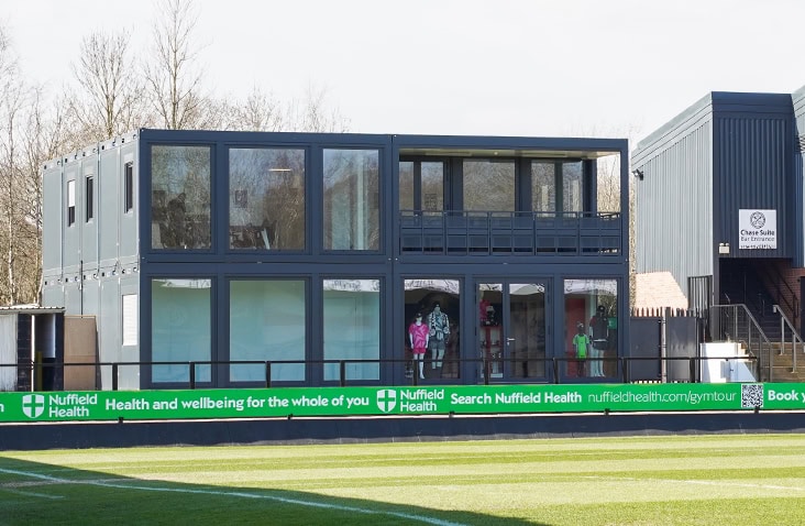 A two-storey modular building constructed from converted shipping containers at a football stadium, featuring large glass panels and balconies overlooking the pitch. 
