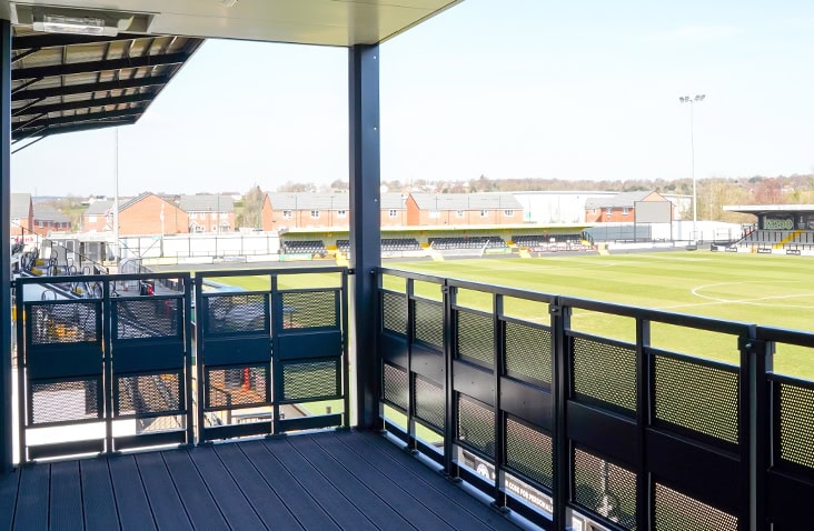 View from the balcony of a modular hospitality building, overlooking the football pitch and stadium seating under a covered roof. 