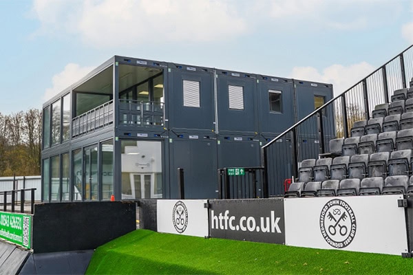 Two-storey modular building constructed from converted shipping containers positioned at a football stadium beside spectator seating.