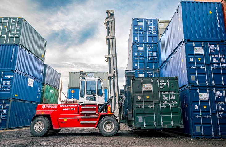 Container handler lifting and positioning a green shipping container between stacked containers at the S Jones Aldridge depot, showing active container handling and yard operations.