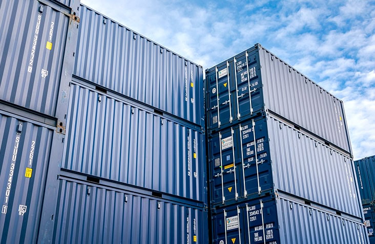 Multiple blue shipping containers are stacked at a container depot under a blue sky, showing secure storage and large-scale container handling operations. 