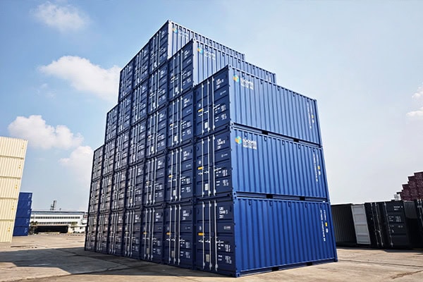 Tall stacks of blue shipping containers arranged in multiple levels at an outdoor storage depot, viewed from below against a blue sky.