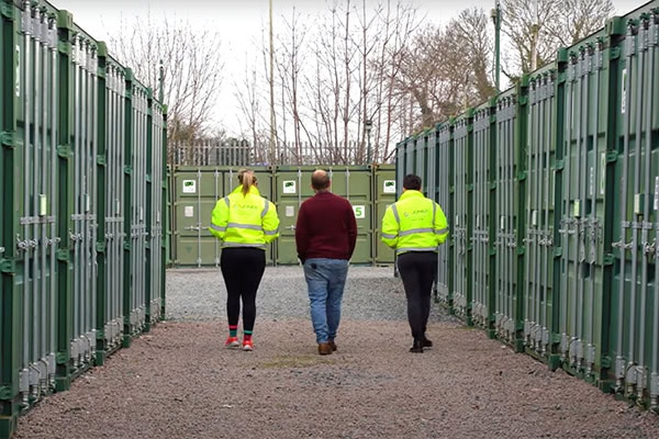 Three people walking between rows of green self storage containers at an outdoor storage facility, with staff in high-visibility jackets.