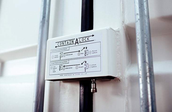 A white Contain-A-Lock protector shroud installed on a white shipping container door. The printed operating instructions are clearly visible on the front, and the metal locking bars run vertically on either side. A padlock shackle protrudes slightly from the bottom of the shroud, showing the lock mechanism in place.