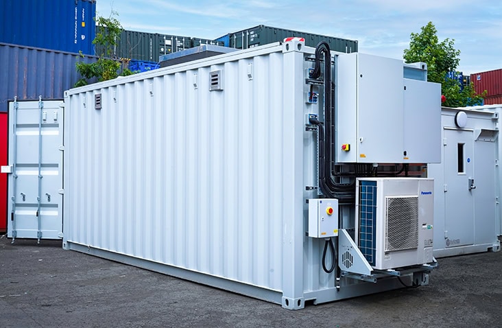 Battery storage container with integrated blast panel and HVAC system, showing a white converted shipping container fitted with external electrical cabinets, ventilation, cooling equipment and safety features for energy storage applications. 