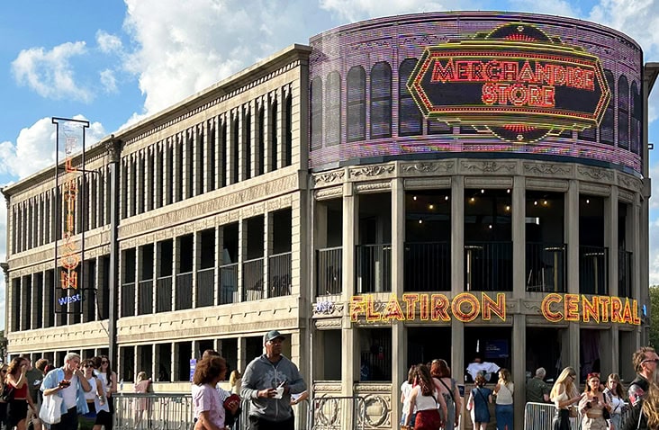 Containerised Flatiron-style building created from stacked shipping containers, featuring a curved corner façade, arched window detailing and illuminated “Merchandise Store” signage at an outdoor event venue.