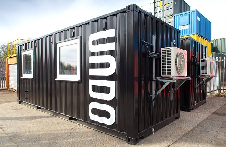 Two containerised mobile laboratories with external HVAC units, showing black converted shipping containers with windows, air conditioning systems and branded exterior, positioned in an industrial yard. 