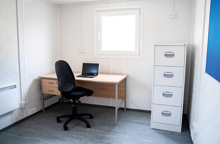 Internal office space inside a site accommodation unit, showing a desk with chair, laptop workstation, filing cabinet, window, and white insulated walls, suitable for temporary site office hire, construction offices, and modular workspace solutions. 