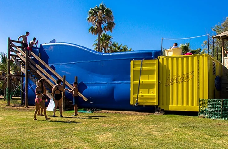 20ft yellow shipping container converted into a water pump enclosure supplying a flow surfing attraction, shown on an outdoor leisure site with inflatable surf ride and access stairs in use.
