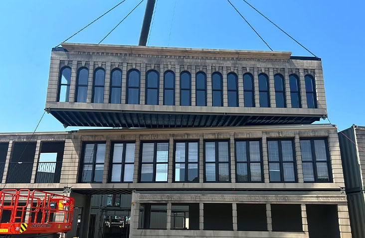 Shipping container building under construction, showing stacked containers being craned into position to form a multi-storey Flatiron-inspired structure with decorative façade panels.