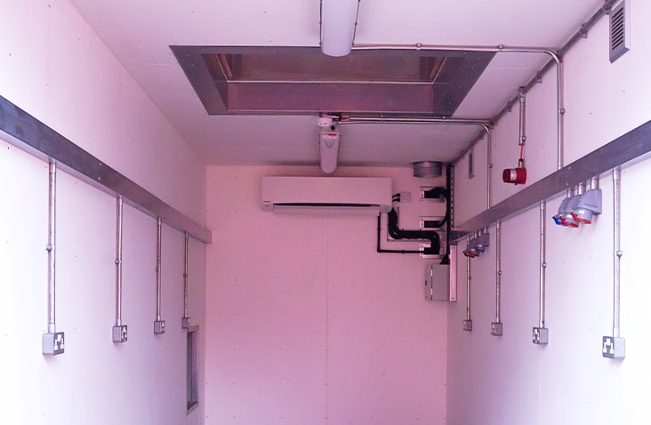 Full internal view of a battery testing container with services on both sides, showing integrated lighting, power outlets, ventilation system, HVAC equipment and safety monitoring infrastructure within a converted shipping container.