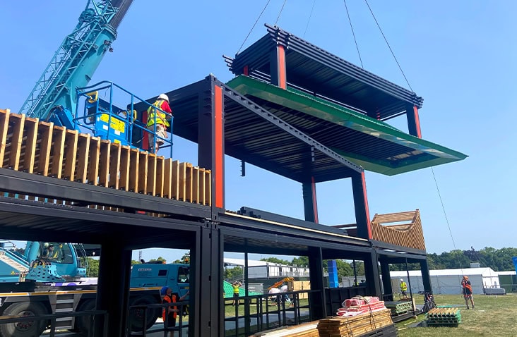 Construction phase of a multi-storey container bar with crane-lifted roof structure, showing stacked shipping containers, steel framework and timber elements during festival venue installation.