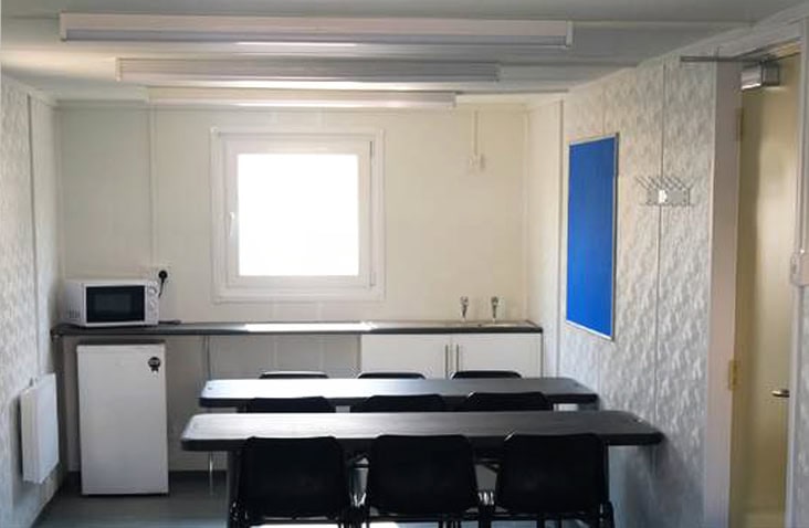Interior of a site canteen accommodation unit, featuring tables and chairs, kitchenette with microwave and fridge, wall-mounted notice board, lighting, and insulated panels, designed for construction site welfare, staff break areas, and canteen unit hire. 