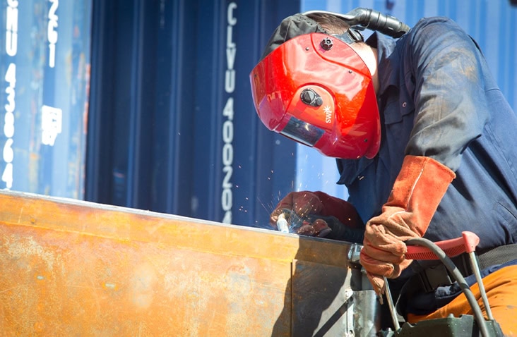 Skilled fabricator welding and modifying a steel shipping container panel, wearing full protective equipment during container fabrication, structural alteration and bespoke container conversion work.