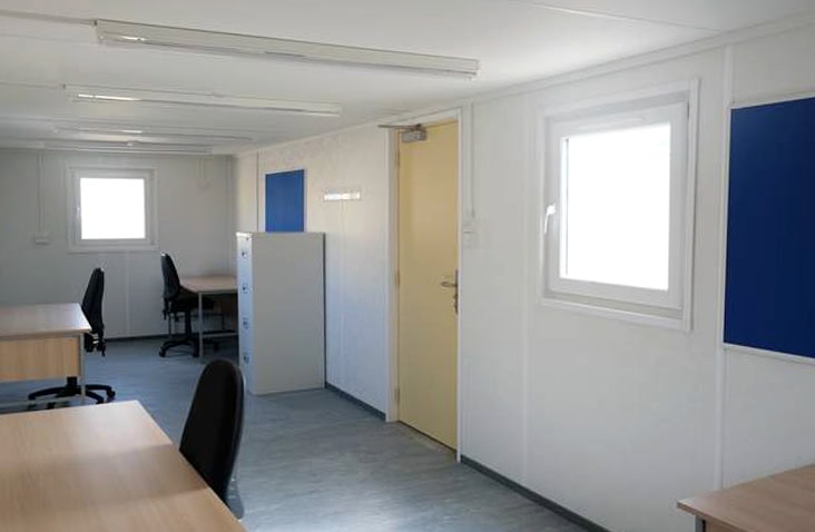     Interior of a white site office with desks, office chairs, filing cabinets and notice boards, featuring windows and fluorescent lighting, designed for temporary office accommodation hire on construction and industrial sites. 
