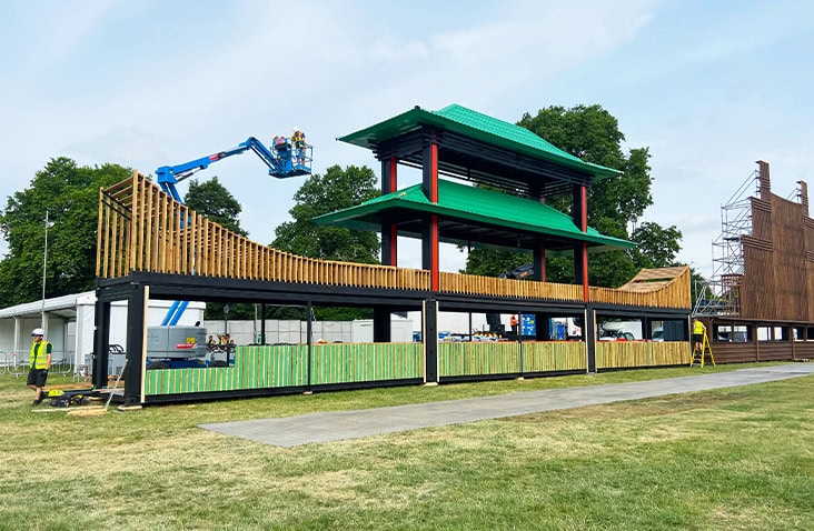 Wide view of a long container bar made from multiple shipping containers, with green roof structure, timber balustrades and open-front serving areas, designed for outdoor event use.