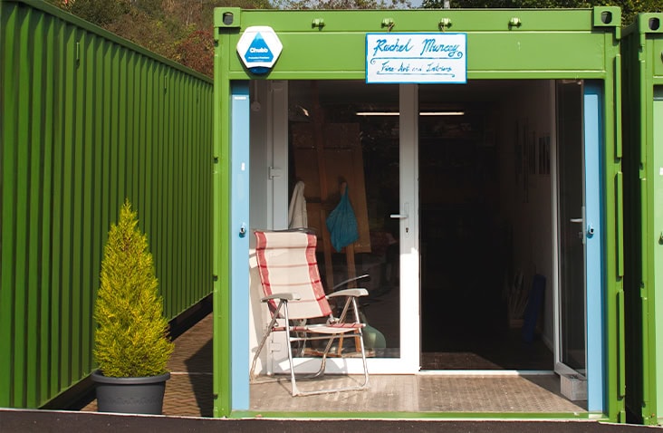 Individual container retail unit, showing a converted shipping container with glazed doors, signage, and a small external seating area used as a creative studio or shop.