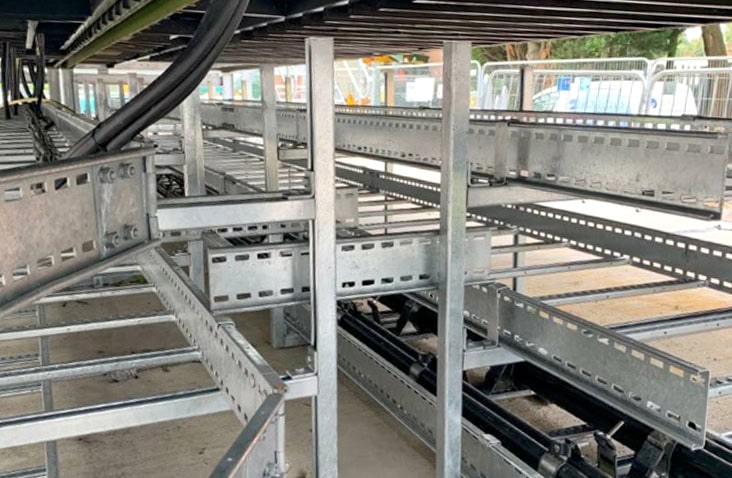 Cable management system beneath containerised switchgear units, showing structured steel cable trays and supports carrying electrical wiring underneath raised containers. 