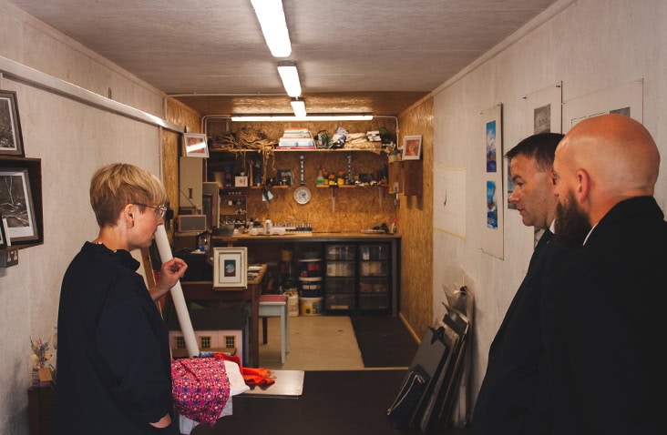 Interior of an insulated container workspace, showing a fitted shipping container studio with lined walls, lighting, storage, artwork displays and people viewing the space as part of a small business unit.