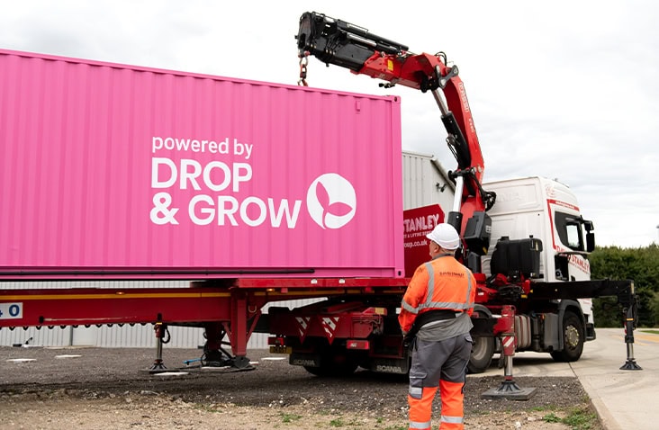 Delivery of a 40ft shipping container vertical farm, showing a crane lorry lifting a branded Drop & Grow container into position during transport and installation at an industrial site. 