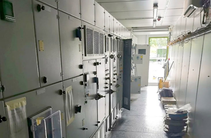 Interior of a containerised switchgear unit, showing installed electrical switchgear panels, control cabinets, cable management, lighting and chequer plate flooring inside a converted shipping container. 
