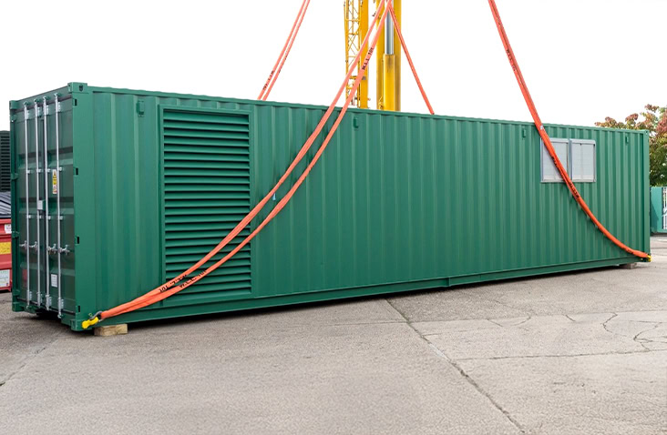 Green battery energy storage container with external ventilation louvres and access doors, secured with lifting slings and positioned on hardstanding, ready for delivery and installation as a containerised energy storage solution.
