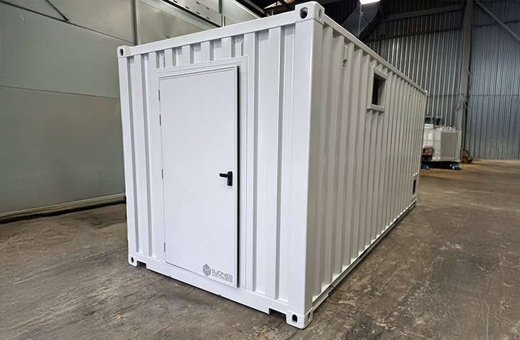 White containerised battery storage unit with personnel access door and external ventilation, shown inside a warehouse prior to delivery, designed for housing battery energy storage systems. 