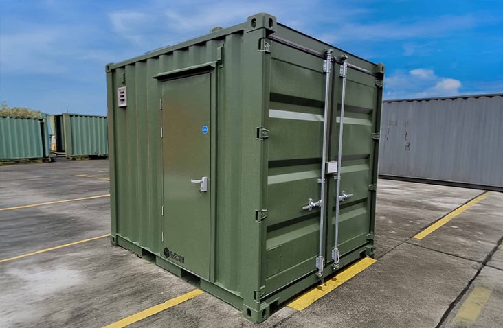 A compact green battery testing storage container is installed at a campus, positioned behind landscaped screening, providing a secure, weatherproof enclosure for lithium-ion battery testing and energy storage. 