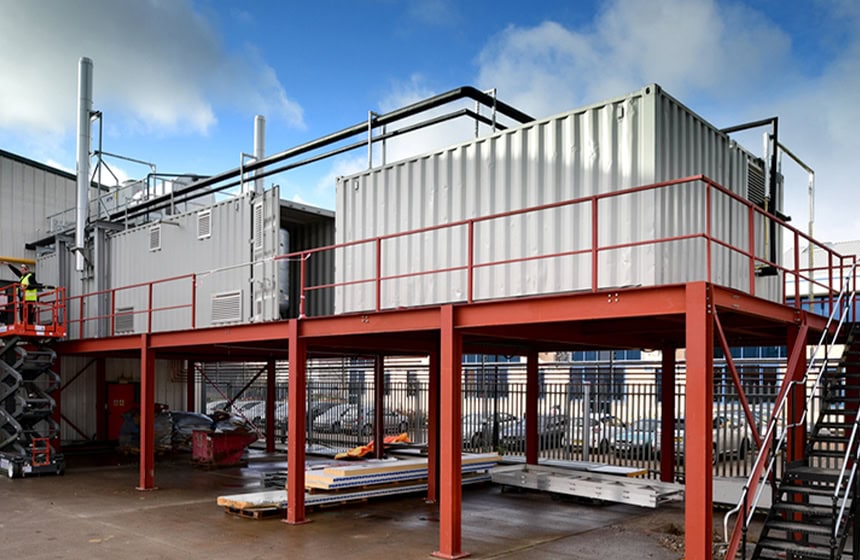 Containerised boiler housing installed on a raised steel platform, showing a white converted shipping container with pipework, ventilation stacks, access stairs and safety railings forming a complete on-site boiler plant solution.
