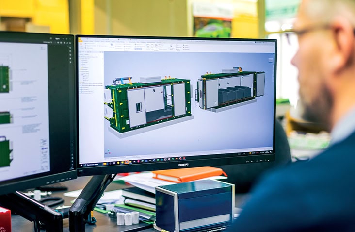 Engineer reviewing a 3D CAD design of a bespoke containerised plant room on a computer screen, showing internal equipment layout and external modifications for an industrial shipping container conversion.